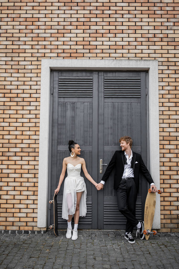 Fröhliche Brautpaare mit Longboard und Skateboard schauen sich in der Nähe des Stadtgebäudes an - Foto, Bild