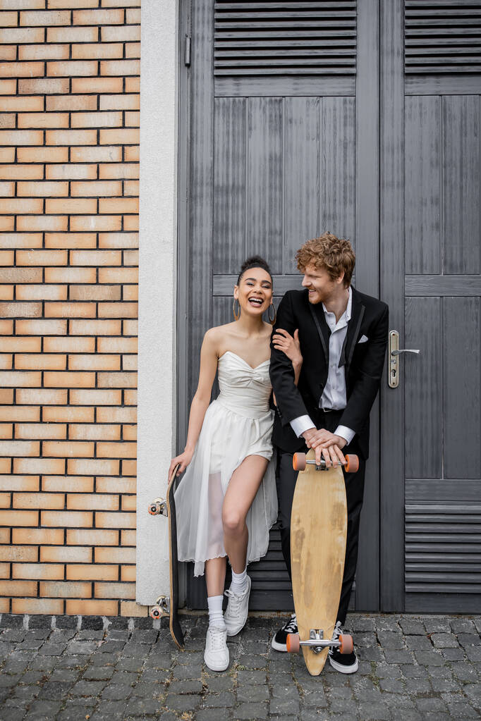 joven pareja interracial con monopatín y longboard cerca de edificio de la ciudad, felicidad, risa - Foto, Imagen