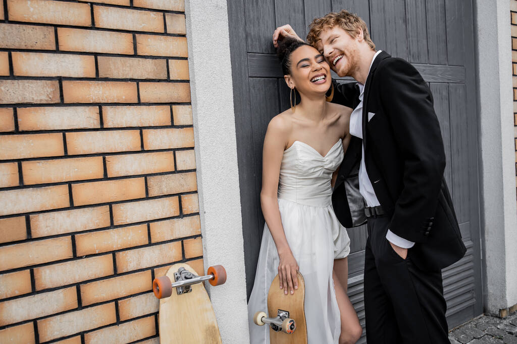 emocionados recién casados multiétnicos riendo cerca de longboard y monopatín en el edificio urbano - Foto, Imagen