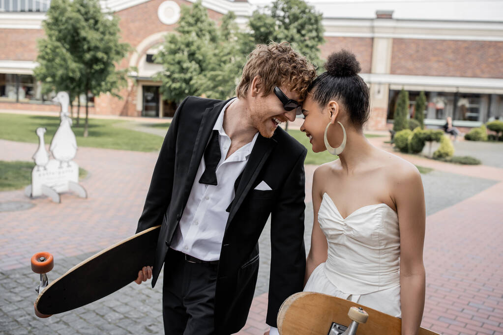 couple interracial avec longboard et skateboard s'amuser dans la rue, tenue de mariage, lunettes de soleil - Photo, image
