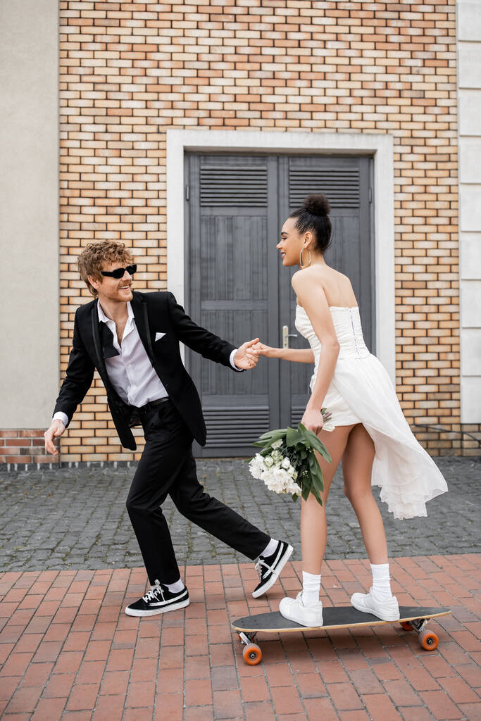 redhead groom in sunglasses holding hands with african american bride with bouquet riding longboard - Photo, Image