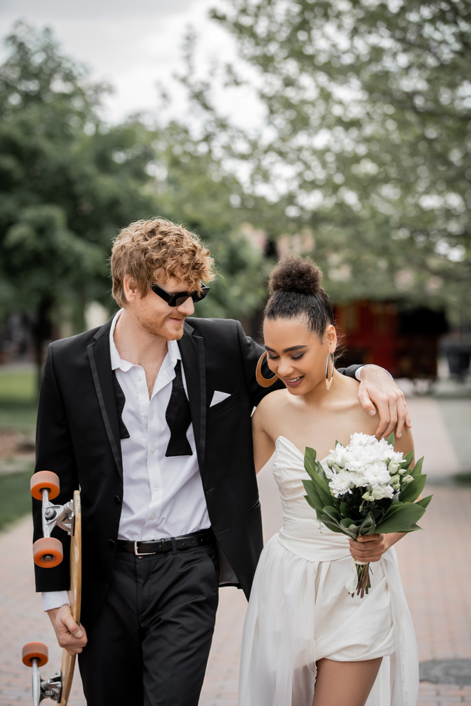 zrzka ženich v slunečních brýlích objímající africké americké nevěsty, chůze s longboard a květiny - Fotografie, Obrázek