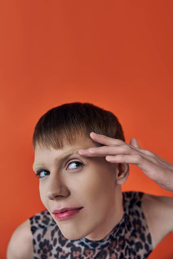 nonbinary person smiling and looking up on orange backdrop, touching eyebrow, queer fashion - Photo, Image