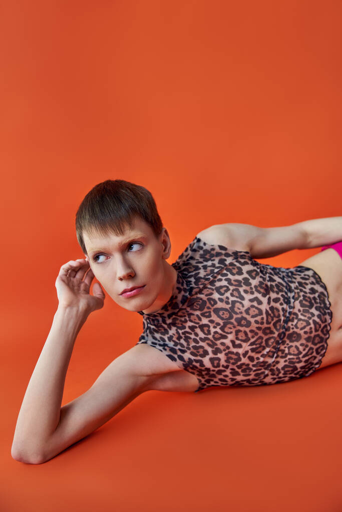 queer person in leopard print sleeveless top lying on orange backdrop, look away, style and fashion - Photo, Image