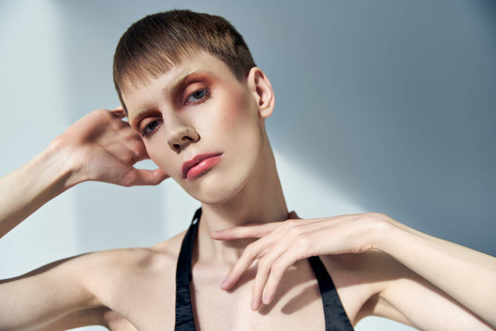 Queer Person mit Make-up posiert auf grauem Hintergrund, Schönheit, Visage, androgynes Modell, Blick in die Kamera - Foto, Bild