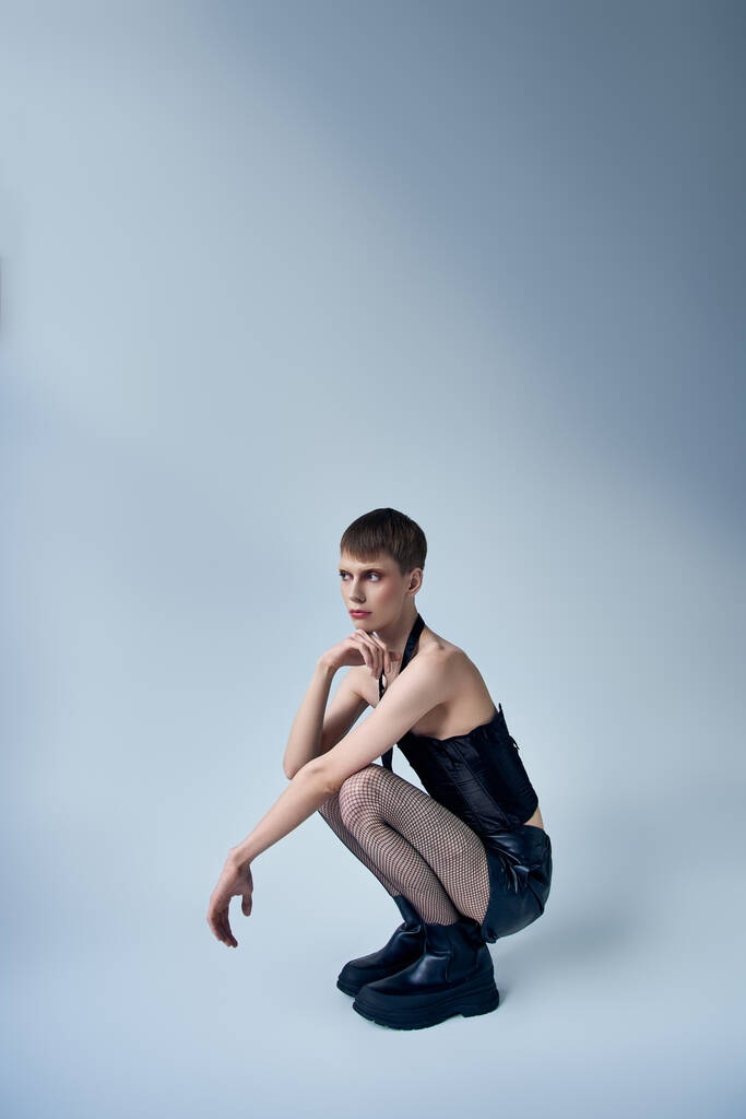 Queer Model in schwarzem Korsett und Netzstrumpfhose auf grauem Hintergrund sitzend, androgyne Person, Mode - Foto, Bild