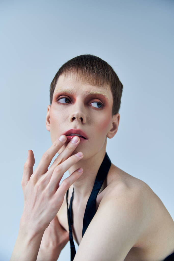 queer model with makeup looking away on grey backdrop, androgynous, touching lip, self expression - Photo, Image