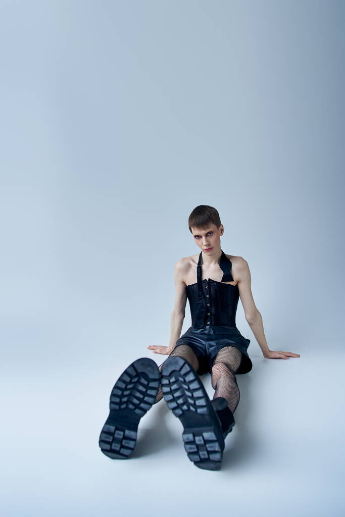 queer model in black corset and fishnet tights sitting on grey, androgynous person, lgbt fashion - Photo, Image
