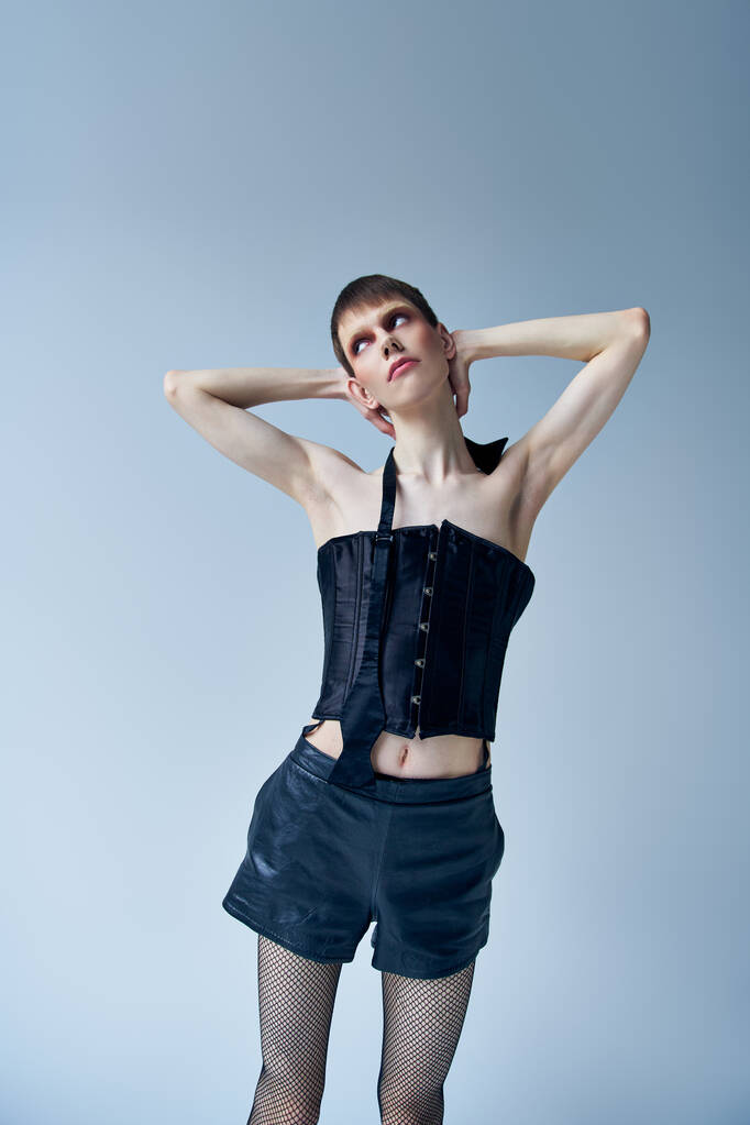 androgynous model in black corset and shorts standing on grey backdrop, lgbt, queer fashion - Photo, Image