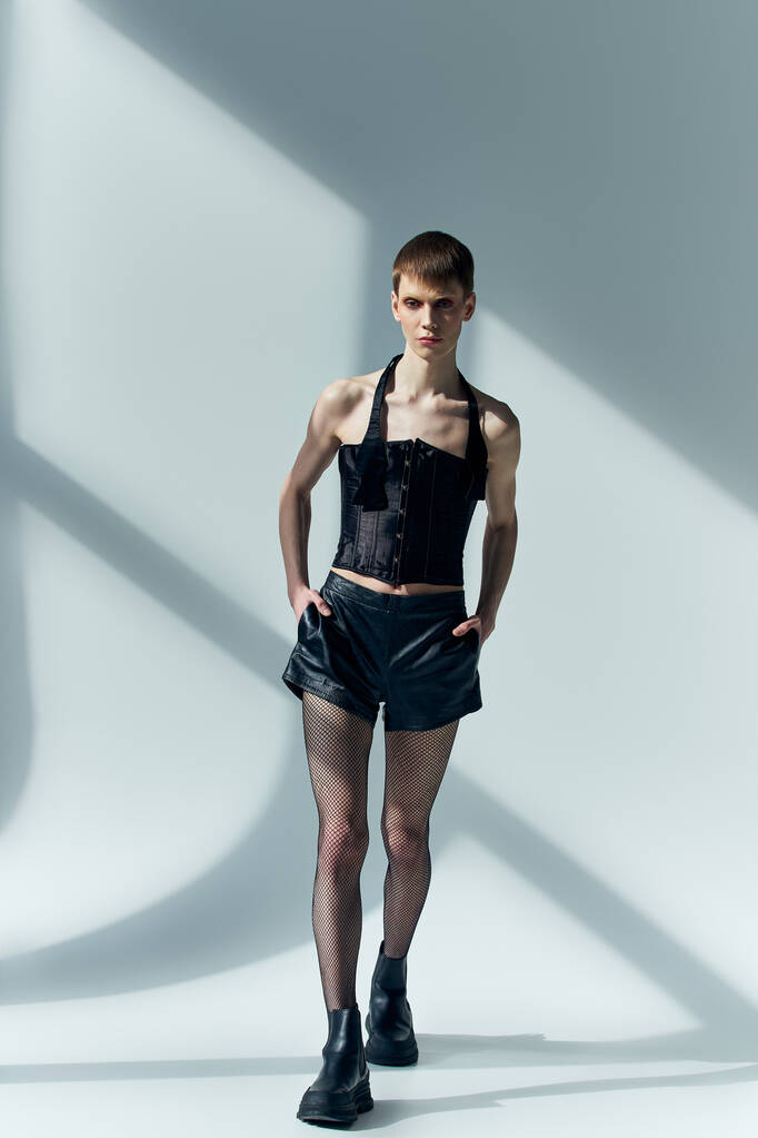 androgynní model v korzetu a černé šortky pózující na šedé, lgbt, podivná móda, ruce v kapsách - Fotografie, Obrázek