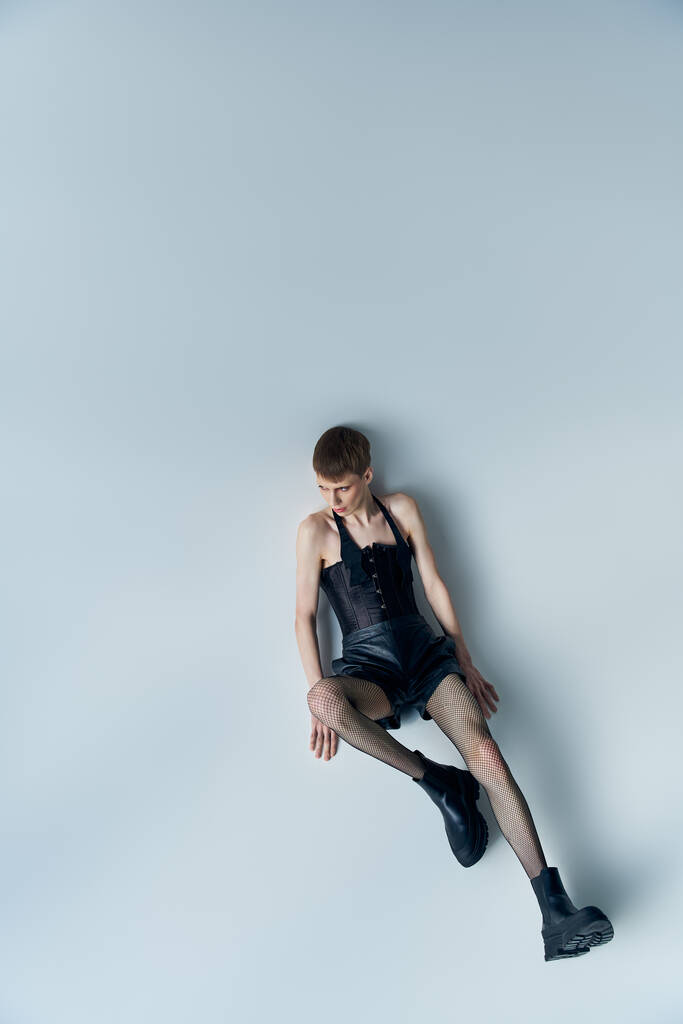 queer model in corset and fishnet tights sitting on grey, lgbt, androgynous style, high angle view - Photo, Image