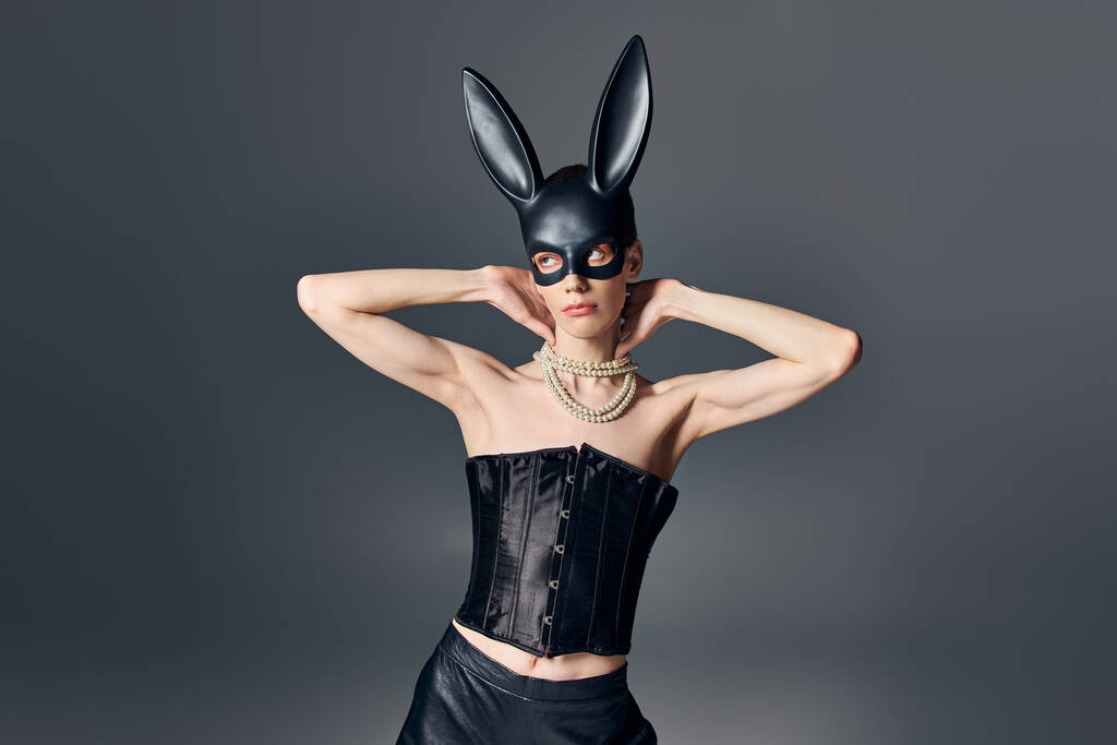 queer model v černém korzetu a bdsm králičí maska na šedé, androgynní, módní, pózovat, dívat se jinam - Fotografie, Obrázek