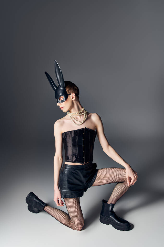 stile audace, persona genderqueer in corsetto posa in maschera coniglietto bdsm su grigio, moda queer - Foto, immagini