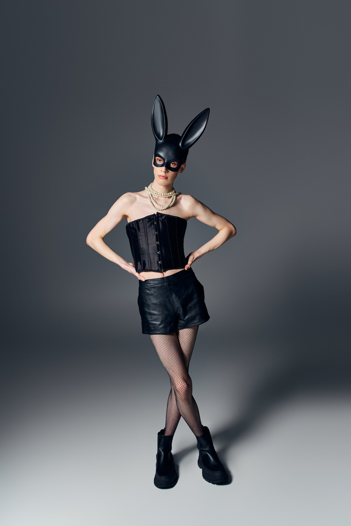 stile, persona genderqueer in corsetto posa in bdsm maschera coniglietto su grigio, moda queer, mani sui fianchi - Foto, immagini