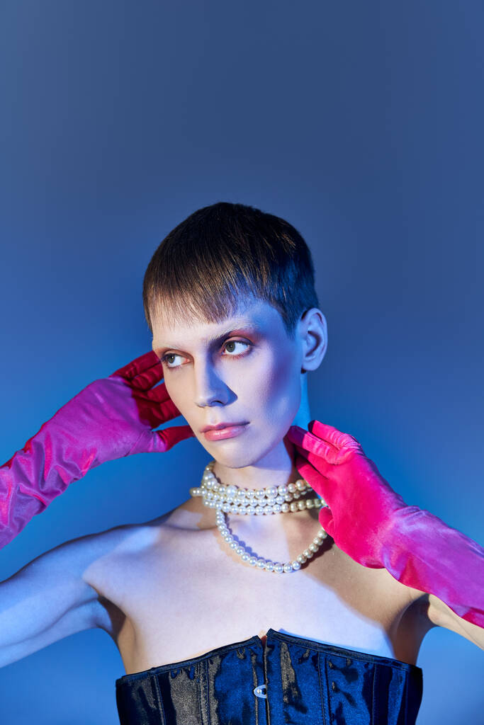 queer persona en corsé negro y collar de perlas posando en guantes de color rosa sobre fondo azul, moda vanguardista - Foto, imagen