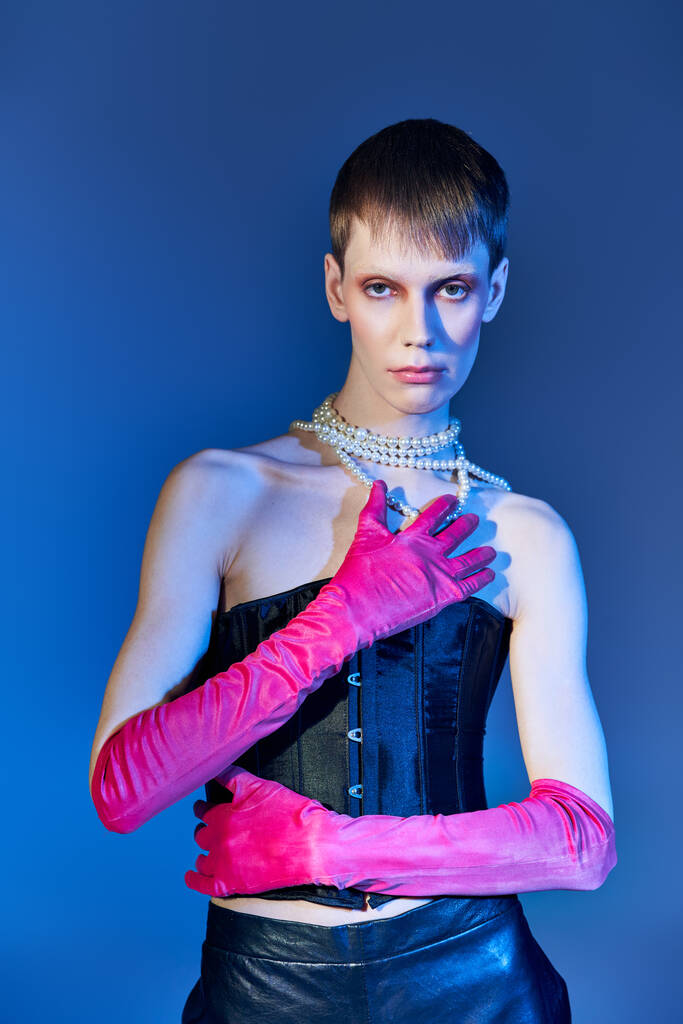 persona no binaria en corsé negro y collar de perlas posando en guantes de color rosa sobre fondo azul, queer - Foto, Imagen