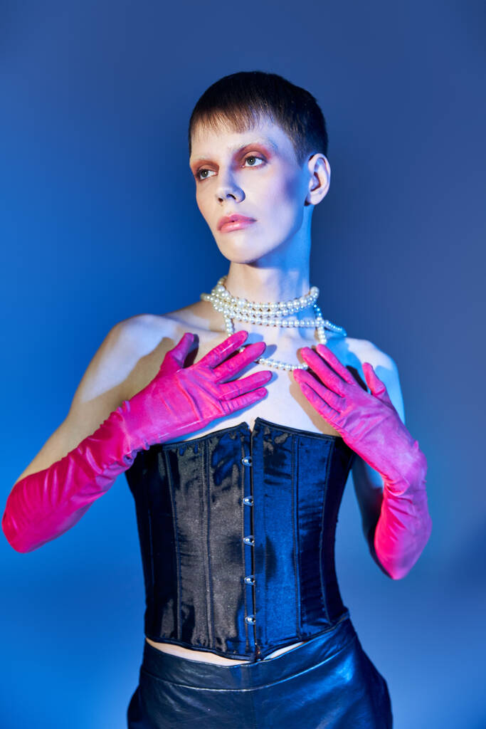 Nichtbinäre Person im Korsett und Perlenkette posiert auf blauem Hintergrund, queeres Modell in rosa Handschuhen - Foto, Bild
