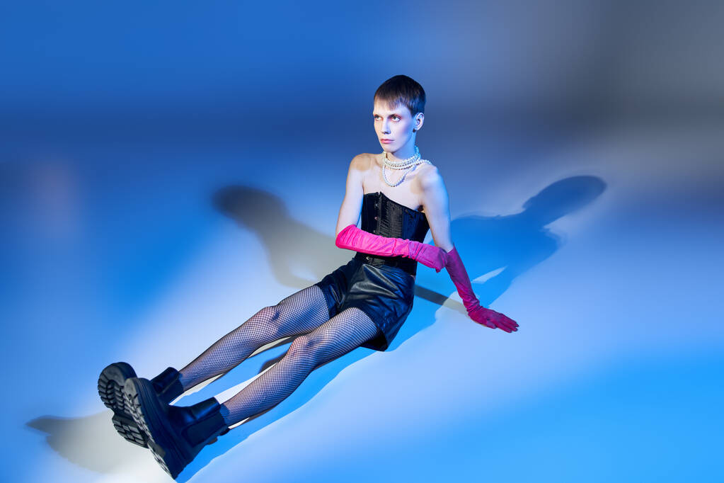 model v korzetu a botách sedící na modrém pozadí, nebinární osoba v růžových rukavicích, teplá móda - Fotografie, Obrázek