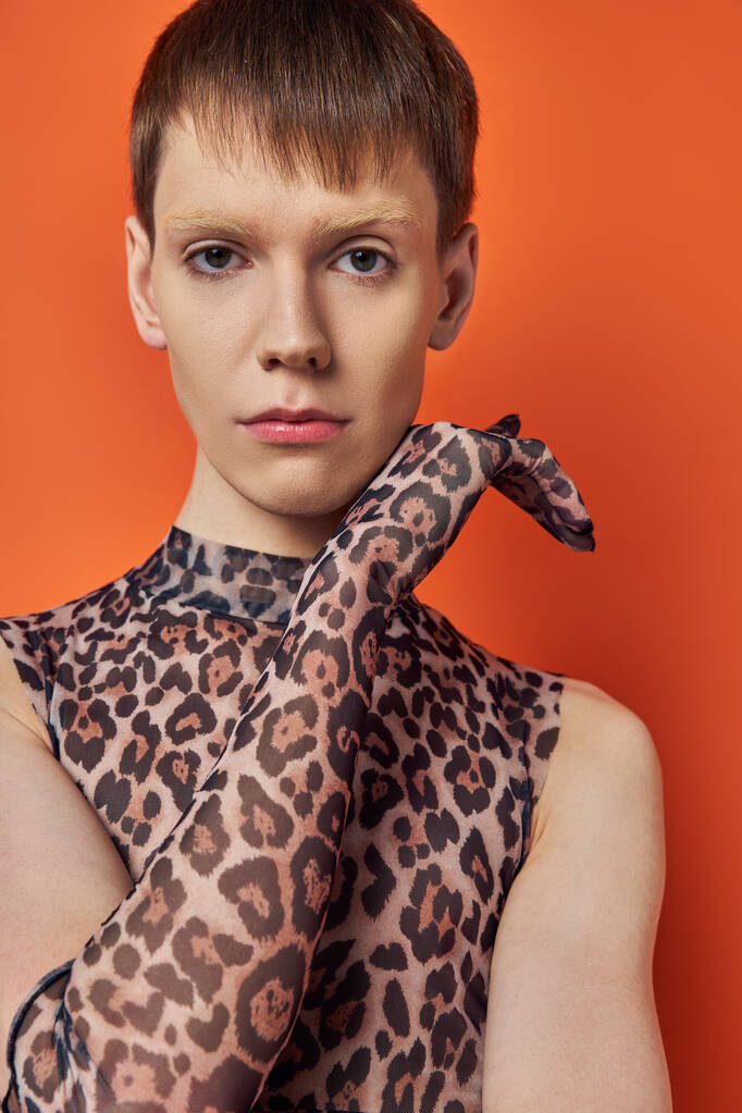 Queer Model im Animal Print Outfit posiert auf orangefarbenem Hintergrund, Genderfluid im Leopardenmuster - Foto, Bild