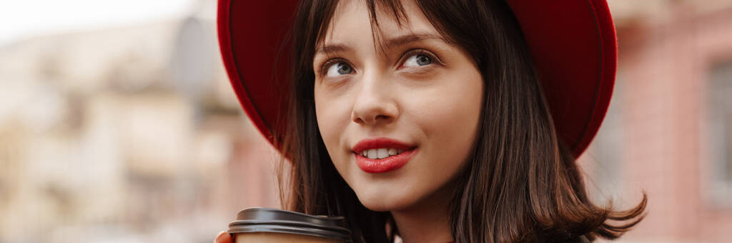 Close up of a smiling young stylish brunette white woman wearing hat and leather jacket with short hair standing on a street holding takeaway cup - Photo, Image
