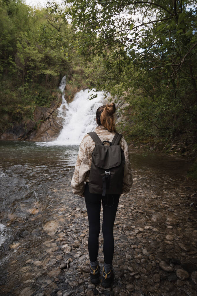 森の真ん中に滝を考えたバックパックとセーターを持つ若い女性 - 写真・画像