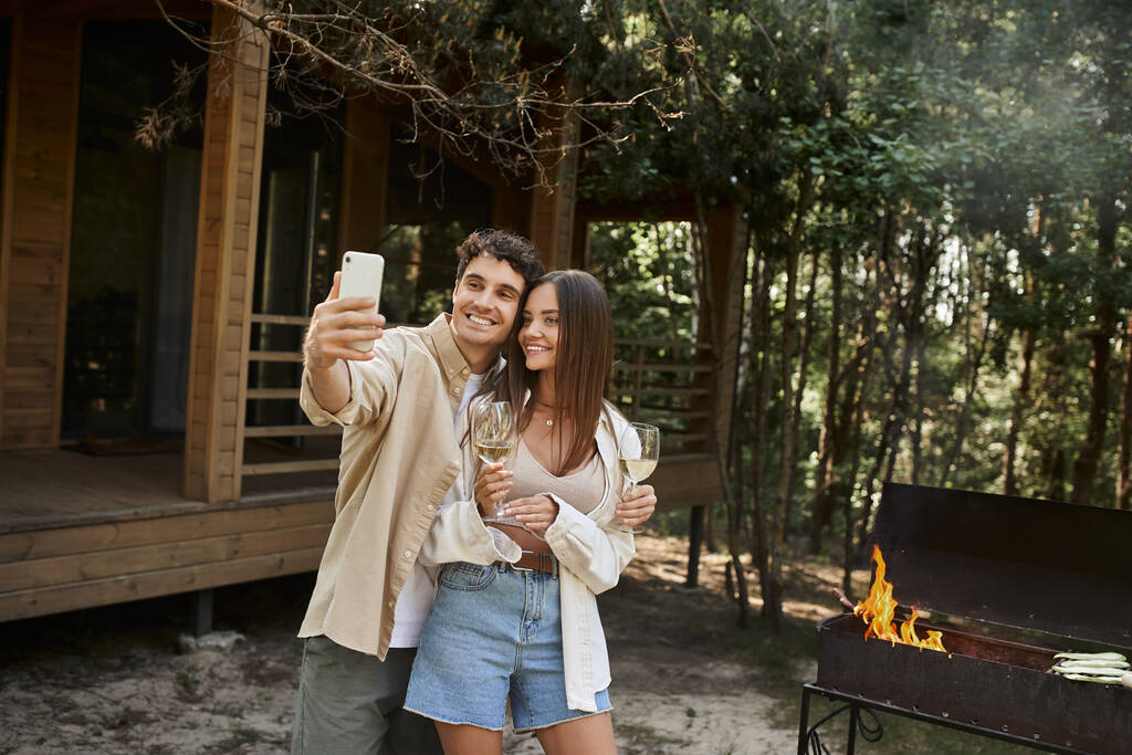 Улыбающаяся пара с селфи на смартфоне рядом с барбекю и домиком отдыха на заднем плане - Фото, изображение