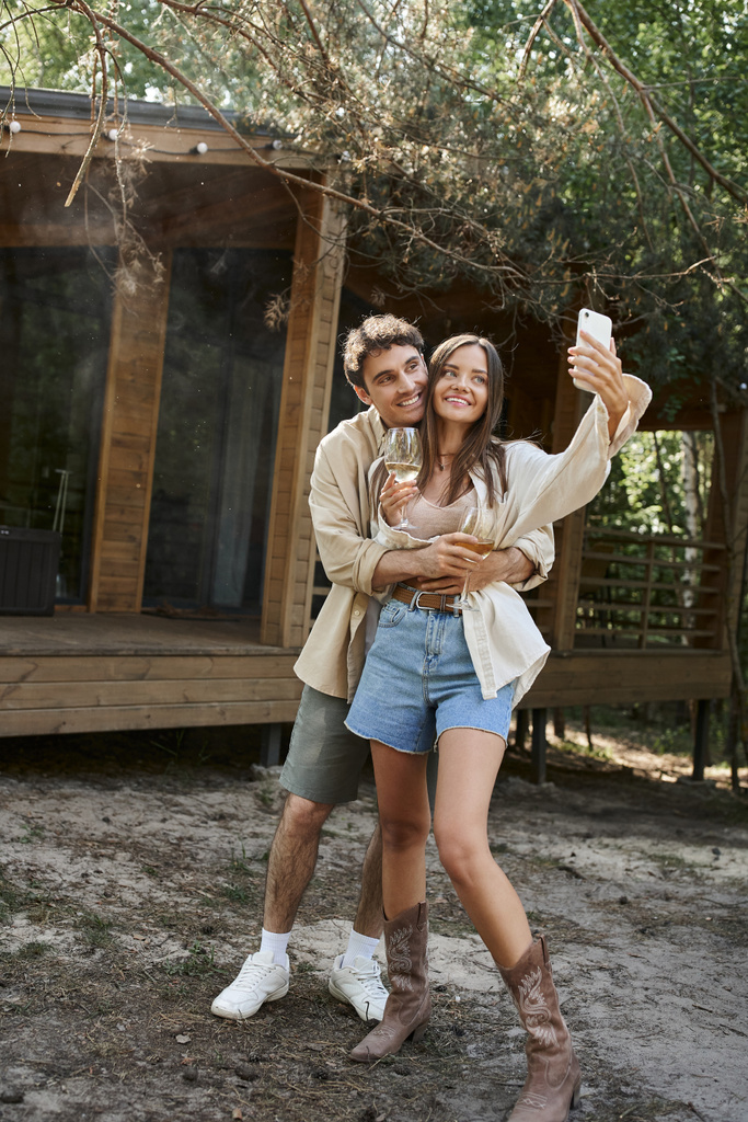 Усміхнений чоловік обіймає дівчину з вином, приймаючи селфі на смартфон біля будинку відпочинку на відкритому повітрі - Фото, зображення