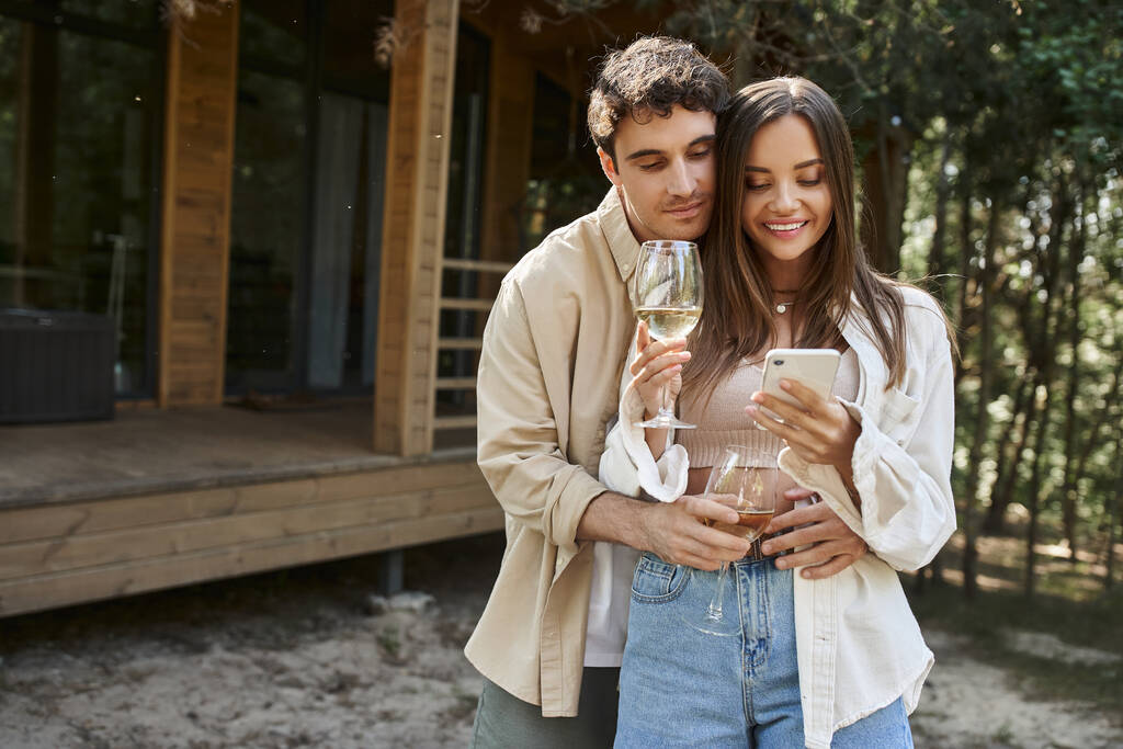 Стильный мужчина обнимает подружку с вином, используя смартфон рядом с домом отдыха на заднем плане - Фото, изображение