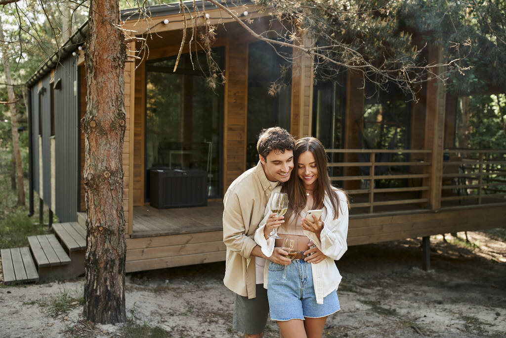Şarabı olan romantik bir çift açık havada yazlık evin yanında akıllı telefon kullanırken gülümsüyor. - Fotoğraf, Görsel