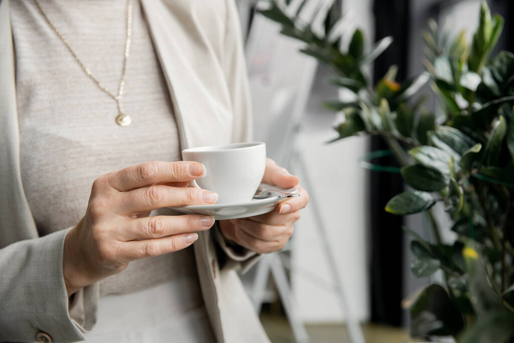vista recortada de la mujer de negocios de mediana edad con estilo de pie con taza de café en la oficina - Foto, imagen