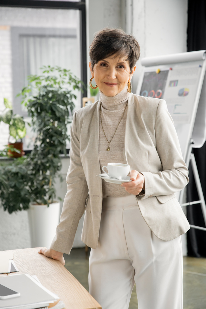 χαρούμενη μεσήλικη επιχειρηματίας με φλιτζάνι καφέ κοντά σε smartphone και έγγραφα στο γραφείο, πορτρέτο - Φωτογραφία, εικόνα