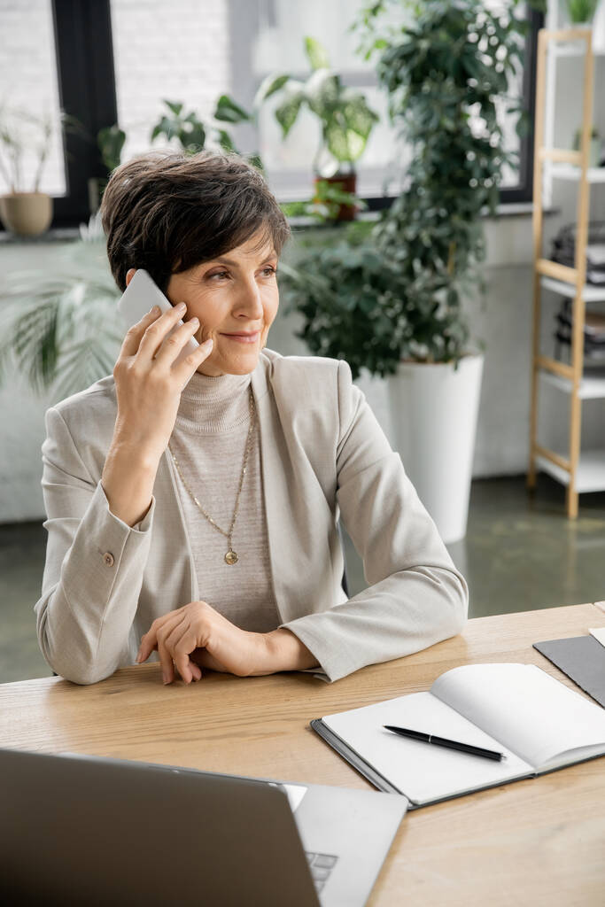 odnoszący sukcesy menedżer korporacyjny w średnim wieku rozmawiający na smartfonie w pobliżu laptopa i notebooka w biurze - Zdjęcie, obraz