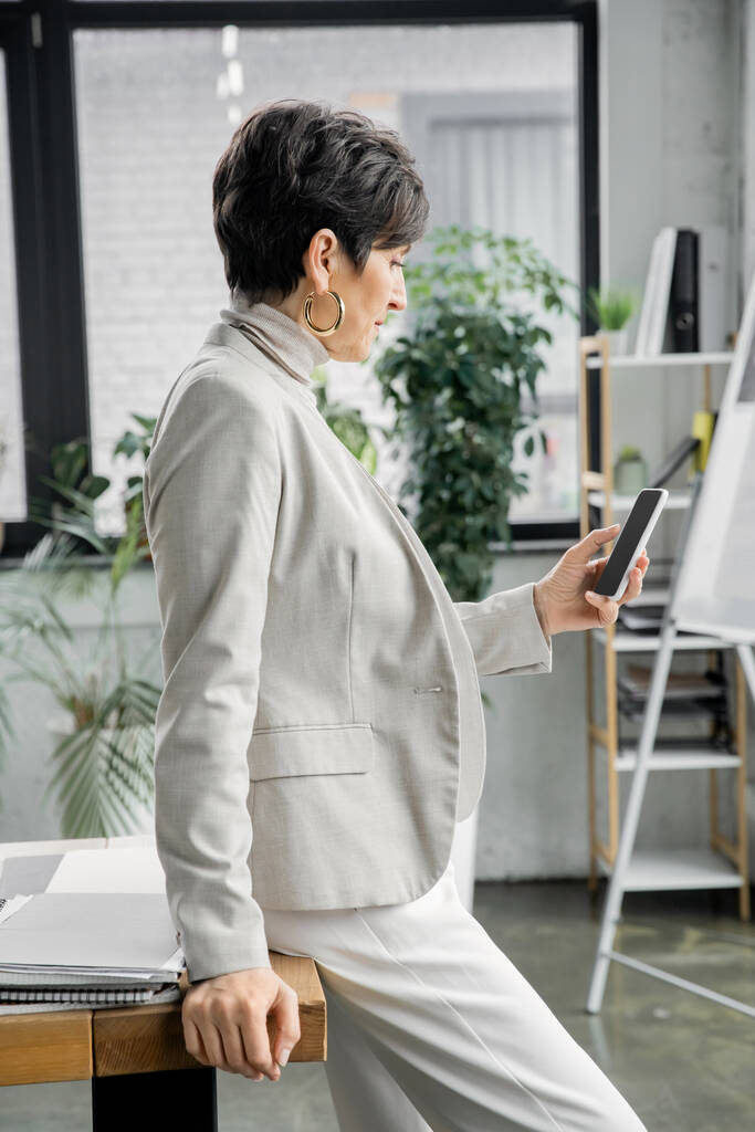 vista lateral de rede de gerente de meia-idade elegante no smartphone perto de documentos na mesa no escritório - Foto, Imagem