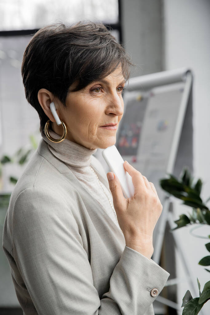 pensivo idade média empresária ouvindo música em fones de ouvido e olhando para longe no escritório - Foto, Imagem