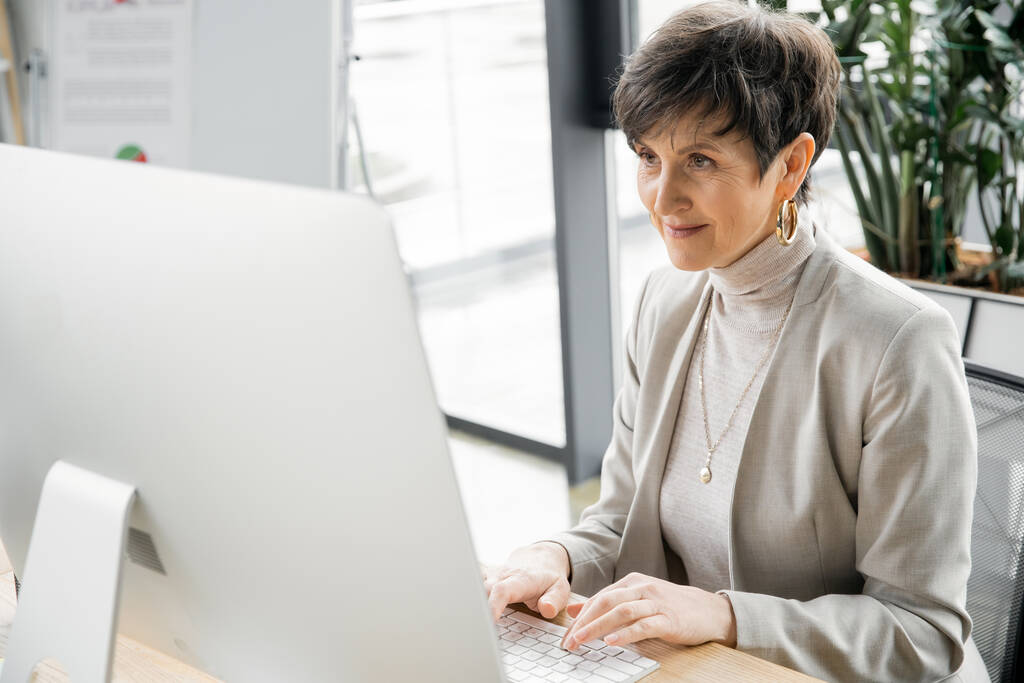 оптимістична бізнес-леді середнього віку друкує на комп'ютері під час роботи в сучасному офісі - Фото, зображення