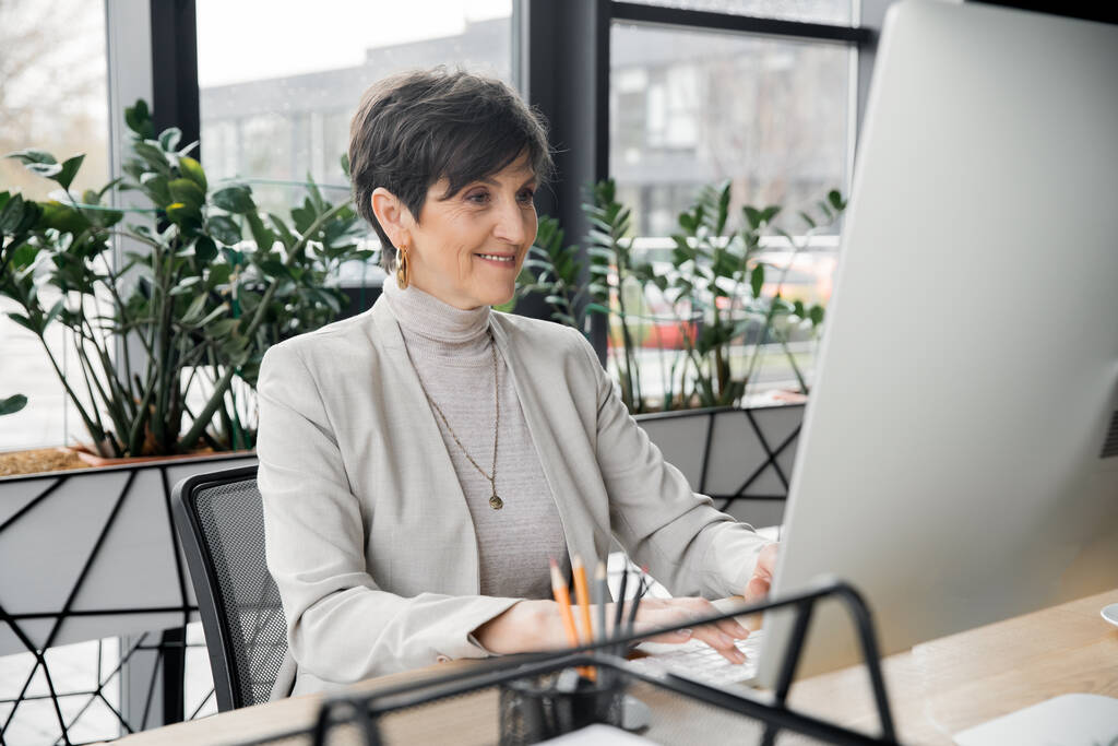 успешный предприниматель средних лет улыбается и работает за компьютером в современном офисе - Фото, изображение