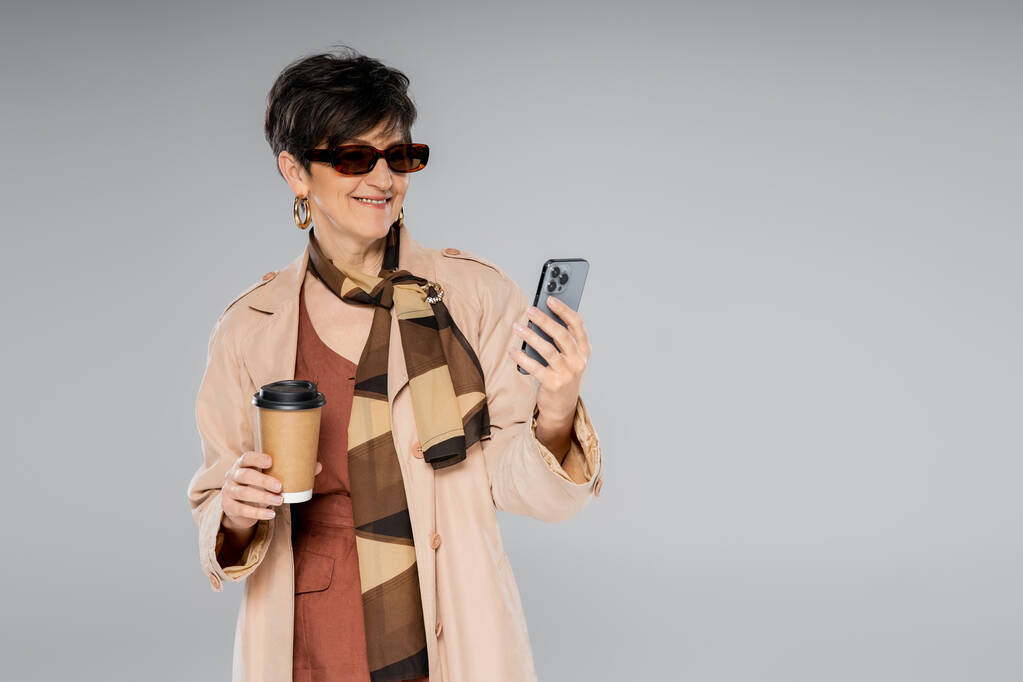 šťastný středního věku podnikatelka s chytrým telefonem a papírovým kelímkem na šedé, podzimní módy - Fotografie, Obrázek
