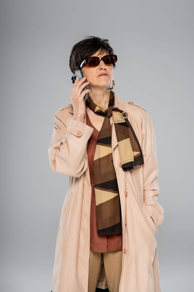 confident and trendy mature businesswoman in sunglasses talking on smartphone on grey, fall style - Photo, Image