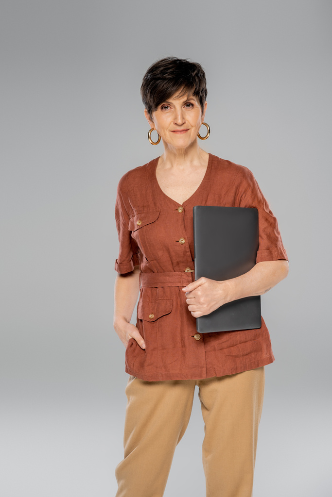 stylowa bizneswoman w średnim wieku z laptopem i ręką w kieszeni na szarej, brązowej kurtce, jesienna moda - Zdjęcie, obraz