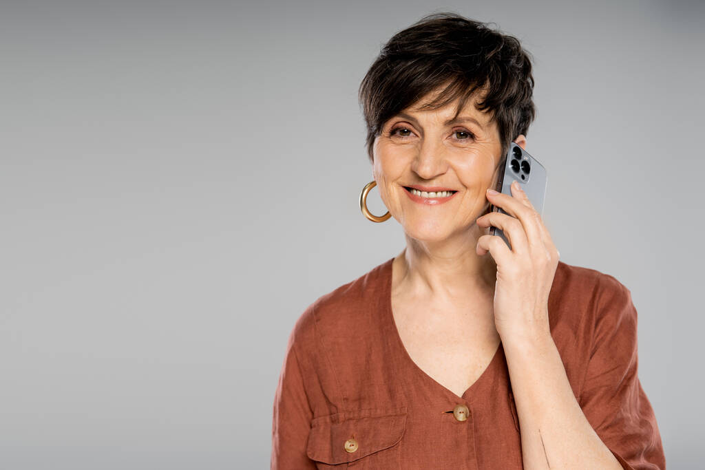 mujer de negocios morena de moda en chaqueta marrón hablando en el teléfono inteligente y sonriendo a la cámara en gris - Foto, imagen