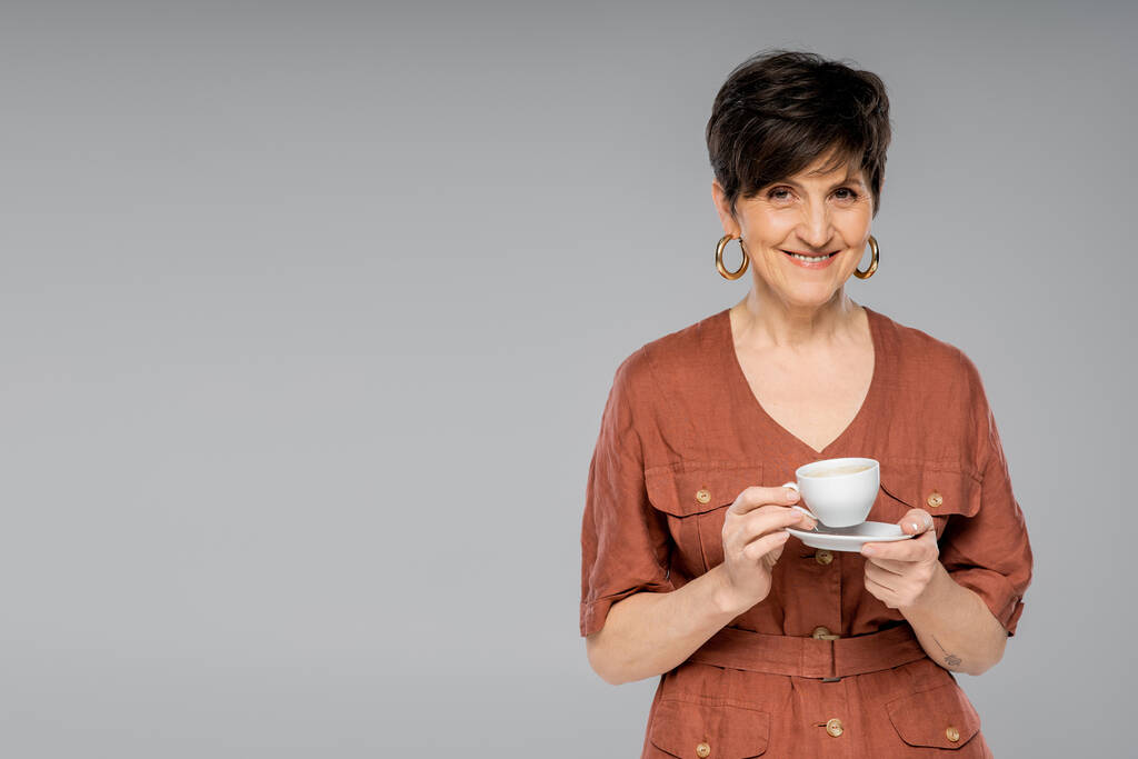 брюнетка середнього віку жінка з чашкою кави посміхається на камеру на сірому, модний вигляд, осінній стиль - Фото, зображення