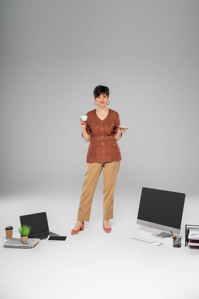 зрелая деловая женщина в стильной осенней одежде, чашке кофе, цифровых устройствах, документах на сером - Фото, изображение