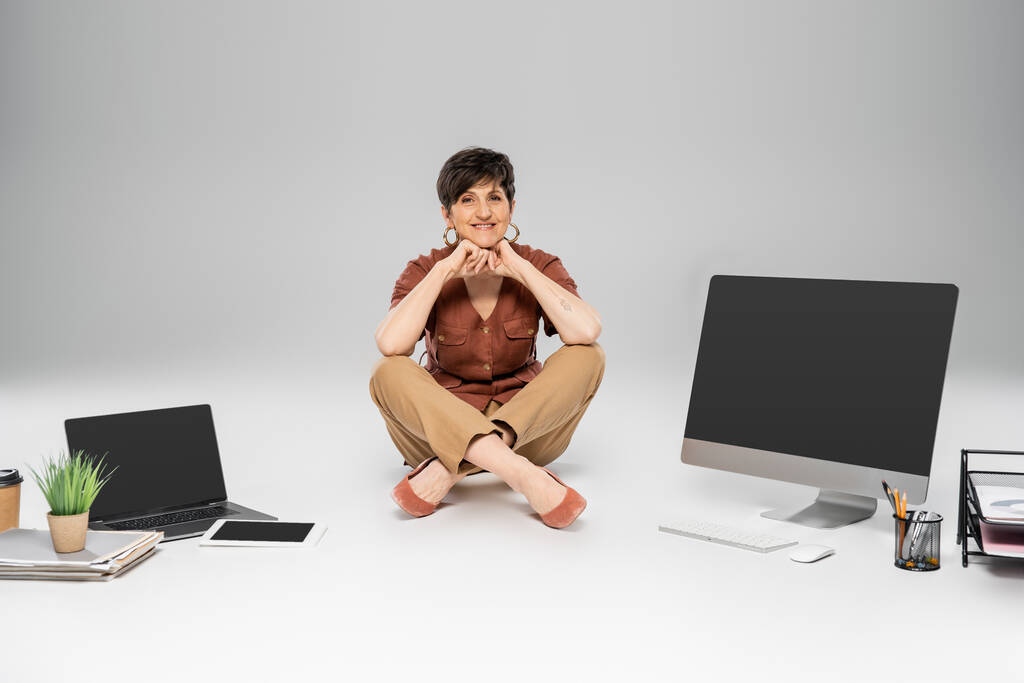 ευτυχισμένη ώριμη επιχειρηματίας κάθεται κοντά σε ψηφιακές συσκευές, έγγραφα, γραφική ύλη σε γκρι, φθινοπωρινή μόδα - Φωτογραφία, εικόνα