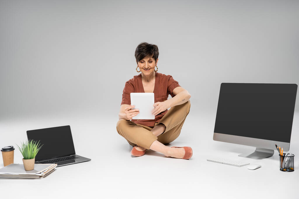 freudige Geschäftsfrau mittleren Alters mit digitalem Tablet in der Nähe von Geräten und Dokumenten auf grau - Foto, Bild