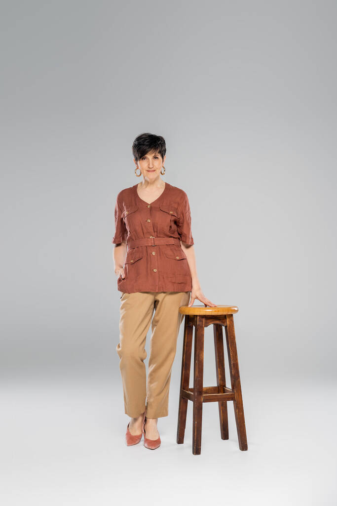 autumn fashion, middle aged woman in trendy clothes near high stool on grey, full length - Photo, Image