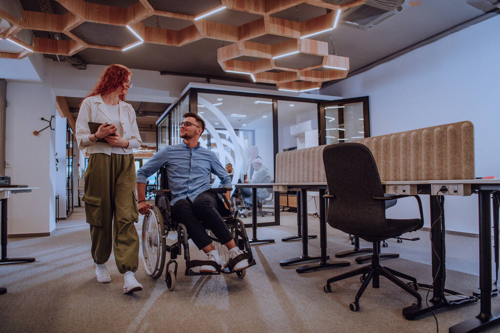 Jovens colegas de negócios, colegas de negócios colaborativos, incluindo uma pessoa em uma cadeira de rodas, passam por um moderno corredor de escritórios de vidro, ilustrando a diversidade, o trabalho em equipe e o empoderamento no - Foto, Imagem
