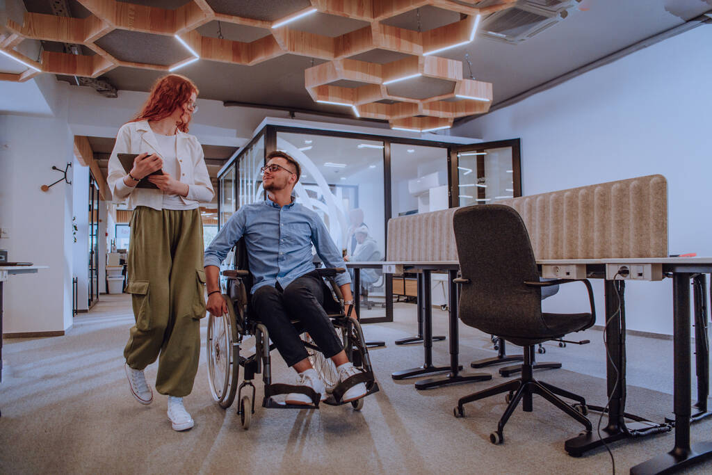Młodzi współpracownicy biznesowi, współpracownicy biznesowi, w tym osoba na wózku inwalidzkim, przechodzą obok nowoczesnego szklanego korytarza biurowego, ilustrując różnorodność, pracę zespołową i upodmiotowienie w - Zdjęcie, obraz