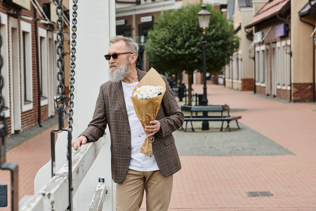 senior man with beard and sunglasses holding bouquet of flowers, urban backdrop, stylish outfit - Photo, Image