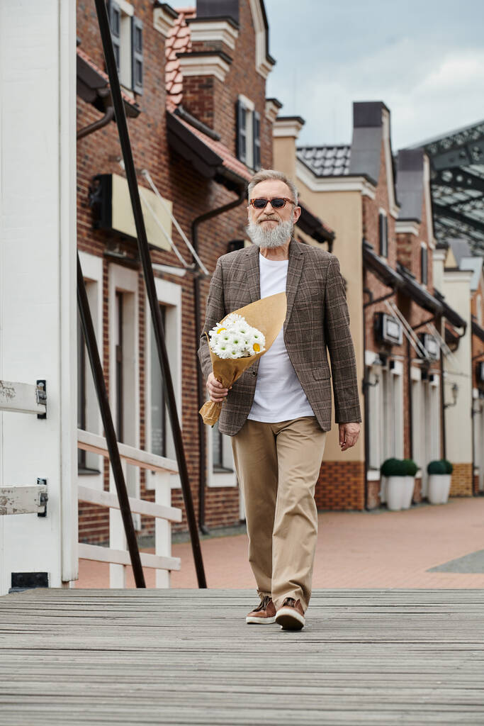 senior man with beard and sunglasses holding bouquet of flowers, walking on urban street, stylish - Photo, Image