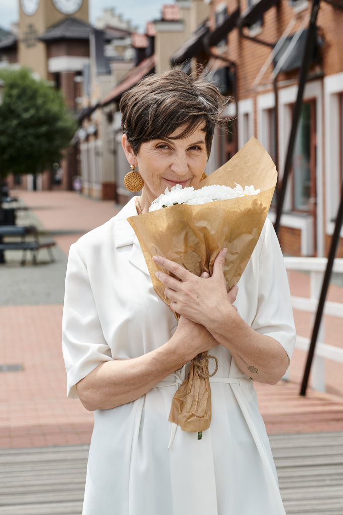 mujer anciana sosteniendo ramo de flores y mirando a la cámara, telón de fondo urbano, verano, traje blanco - Foto, Imagen